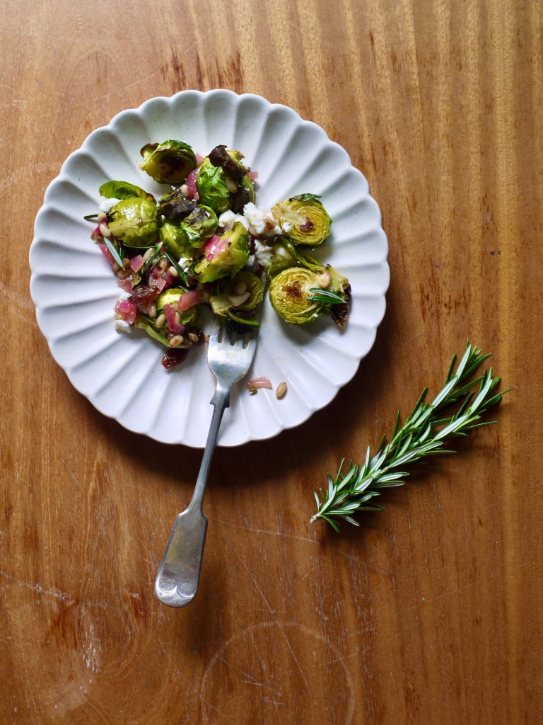 Roasted brussels sprouts on a plate with a fork, with a sprig of rosemary next to it on the table. This image links through to a web page with the recipe.