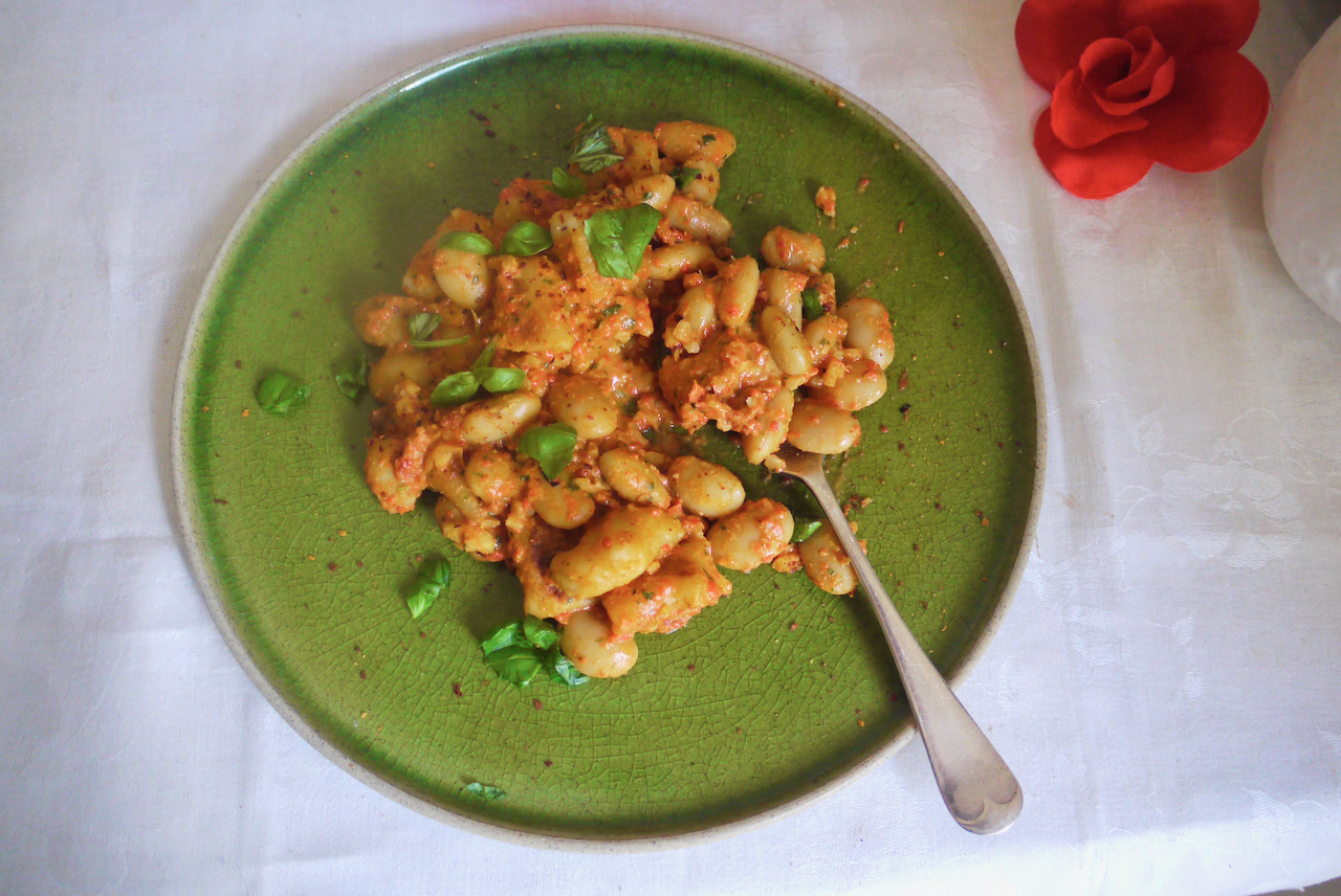 A green plate of gnocchi with a fork rests on a white tablecloth with a red fabric rose nearby.
