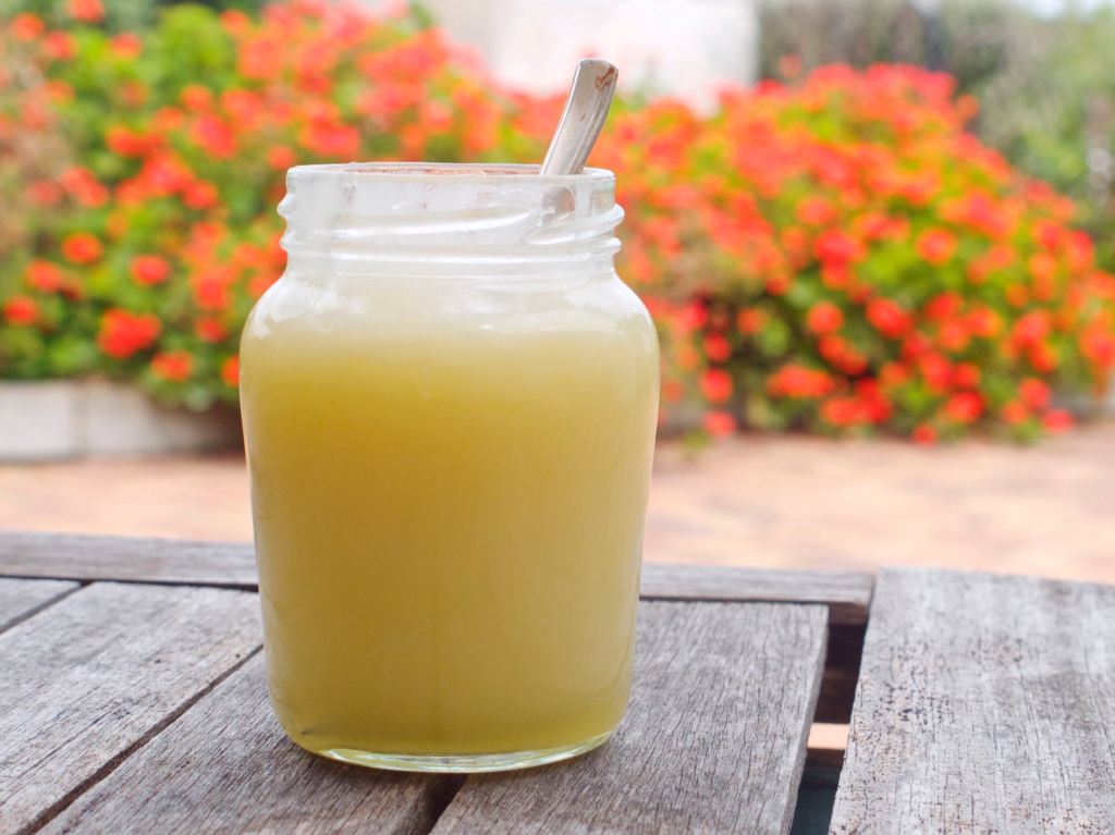 A jar of lemon curd with a spoon in it, on a table, with red geraniums visible in the background. This image links through to a web page with the recipe.