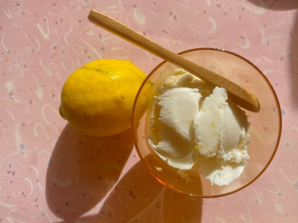 No-churn lemon ice cream in a bowl on a pink background with a lemon. This image links through to a web page with the recipe.