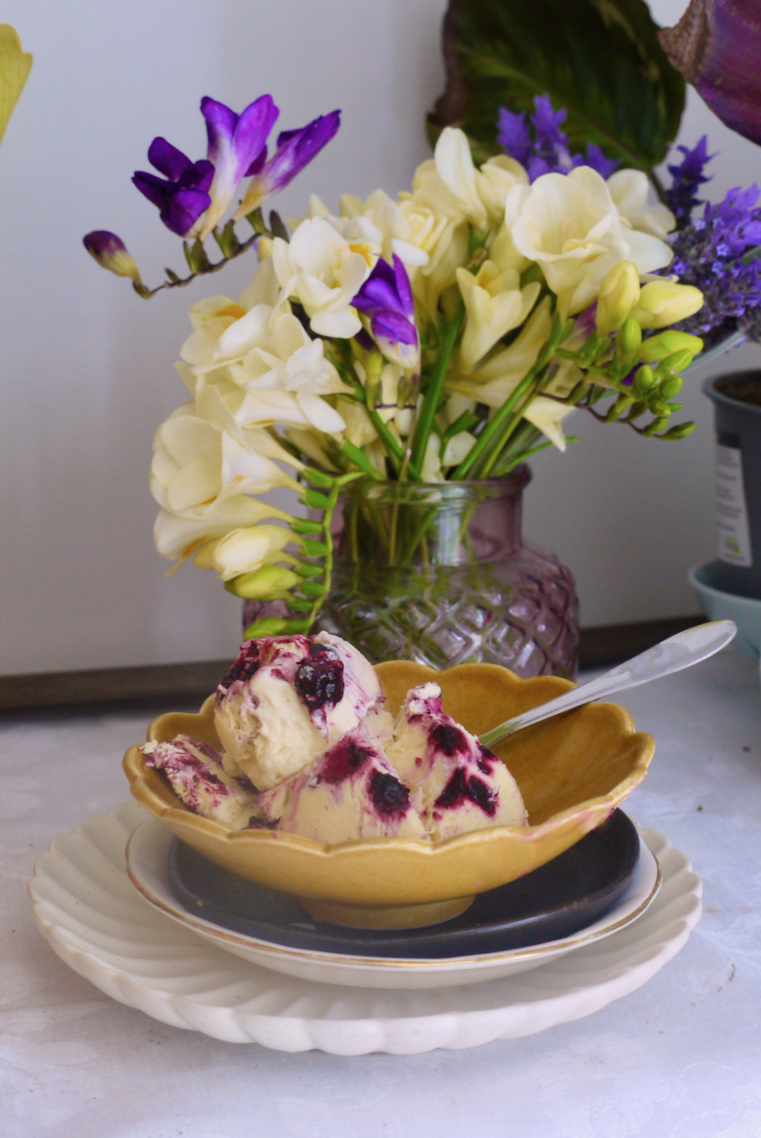 a brown scalloped bowl of blueberry ice cream on a pile of stacked place with a vase of freesias in the background