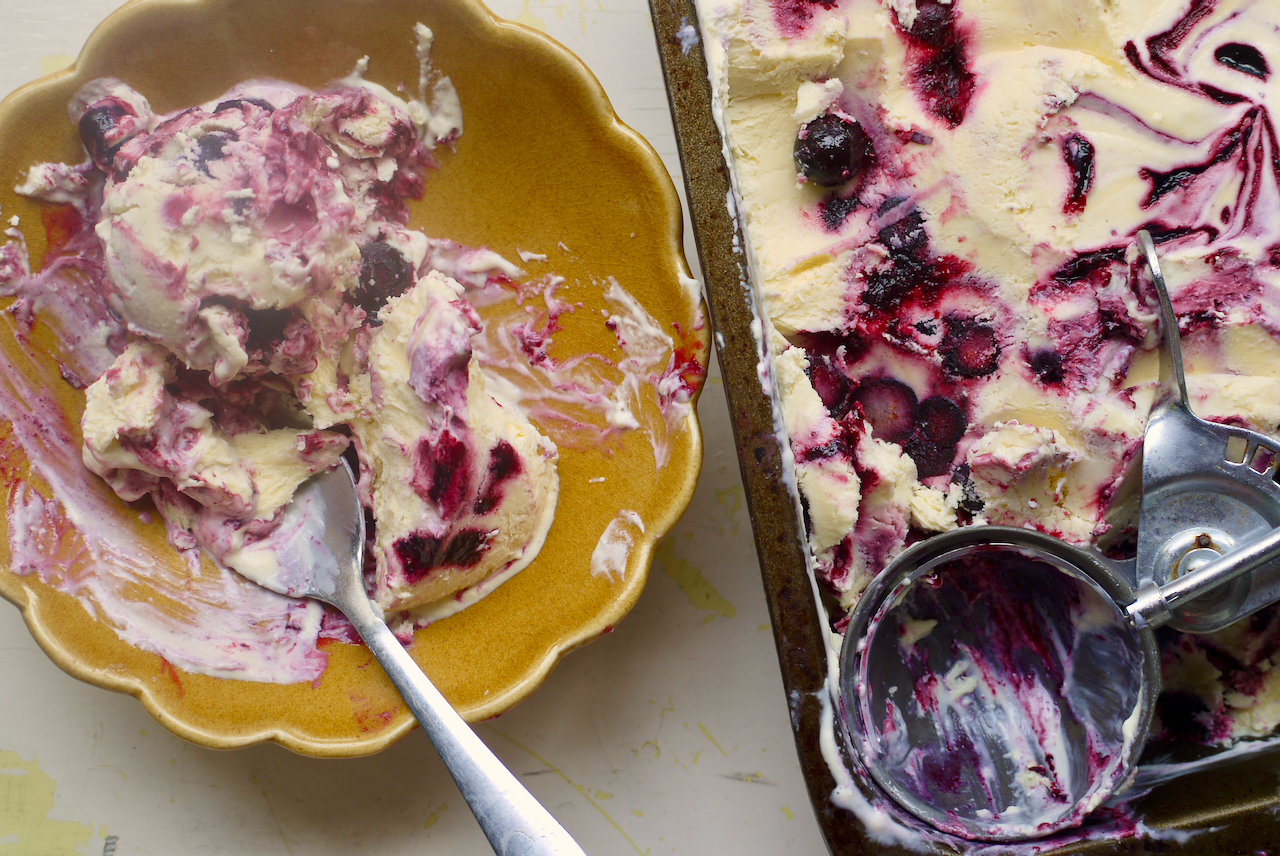 A brown scalloped bowl of blueberry ice cream, partially eaten, next to a tin of ice cream