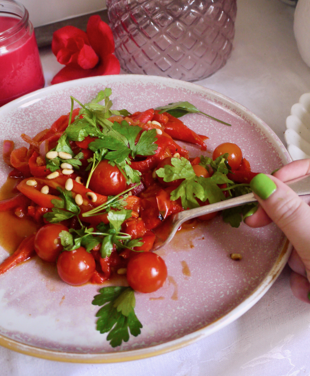 A hand with green fingers, using a fork to get some piperade from a pink plate. In the background is a pink glass vase and a red rose.