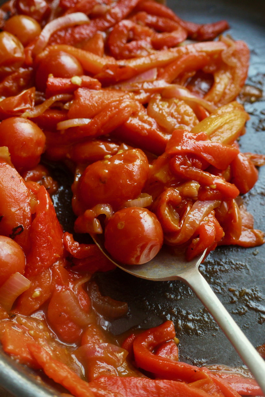 A serving spoon lifting a spoonful of piperade from a frying pan