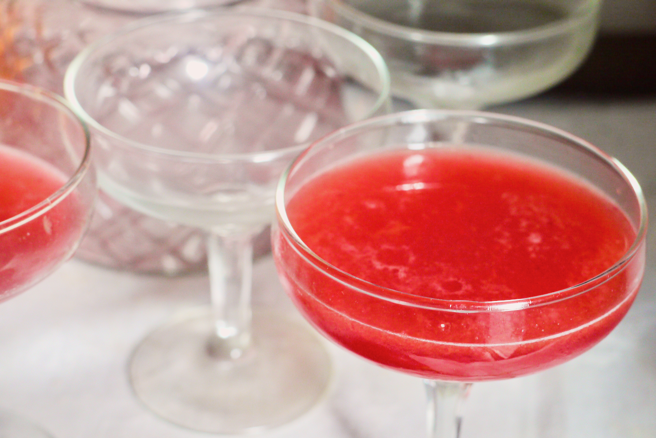 A close up of a tamarillo cocktail with empty cocktail glasses in the background