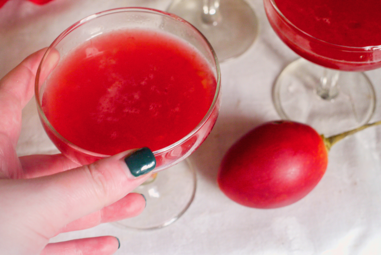 A hand with dark green fingernails picking up a tamarillo cocktail, with a tamarillo beside it on a white tablecloth