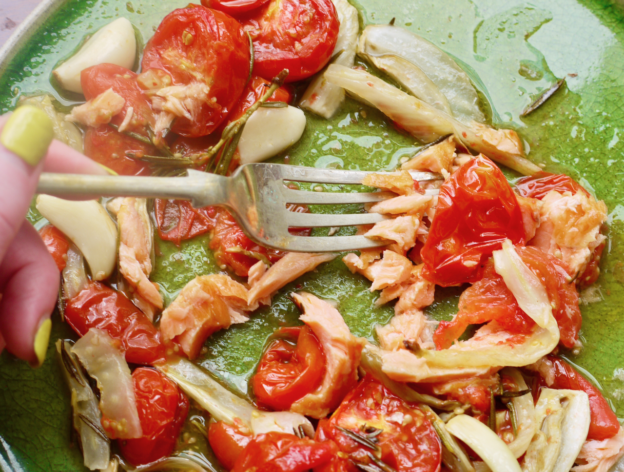 A hand with green painted nails using a fork to spear a piece of salmon on a green plate with cherry tomatoes and fennel also on it