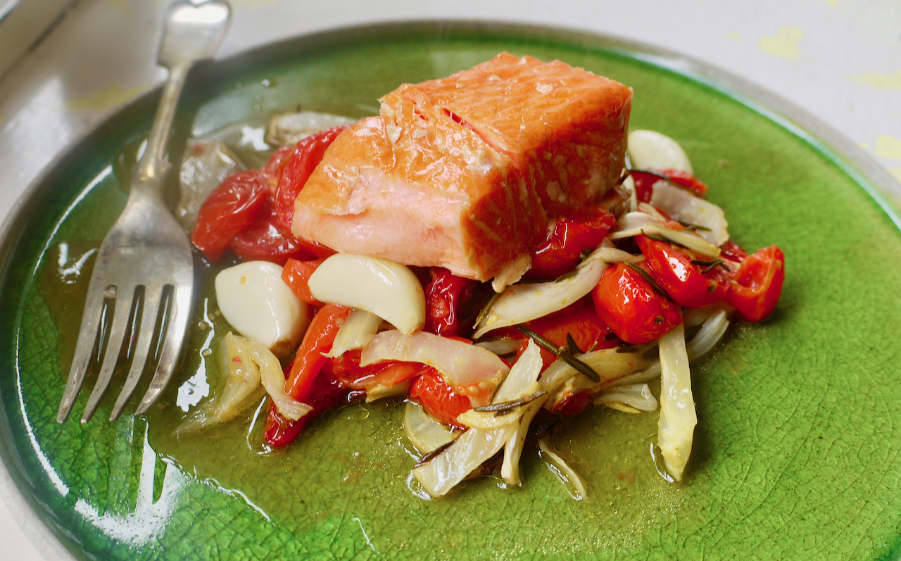 A piece of salmon resting on roasted cherry tomatoes and fennel on a green plate with a fork