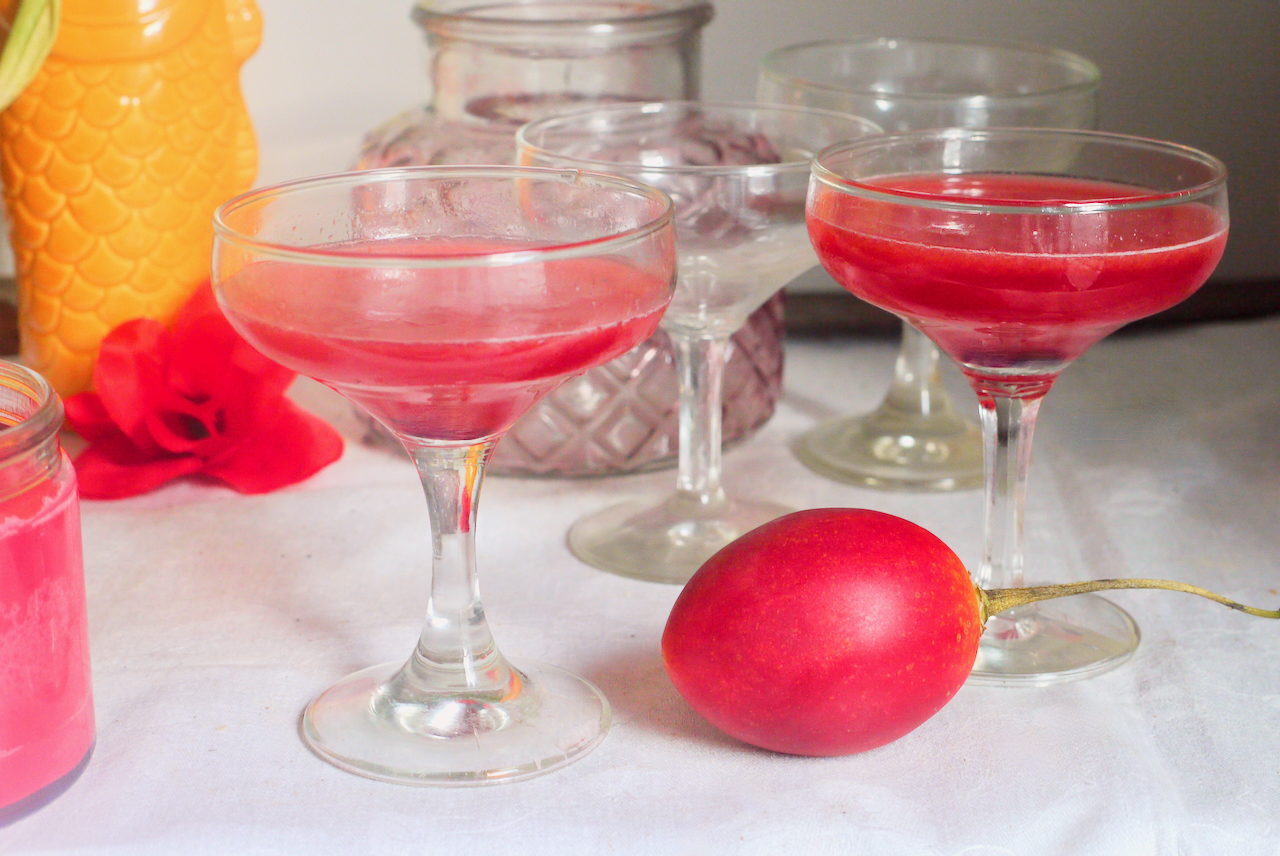 Two tamarillo cocktails on a white tablecloth with a tamarillo in front of them and a red fabric rose in the background