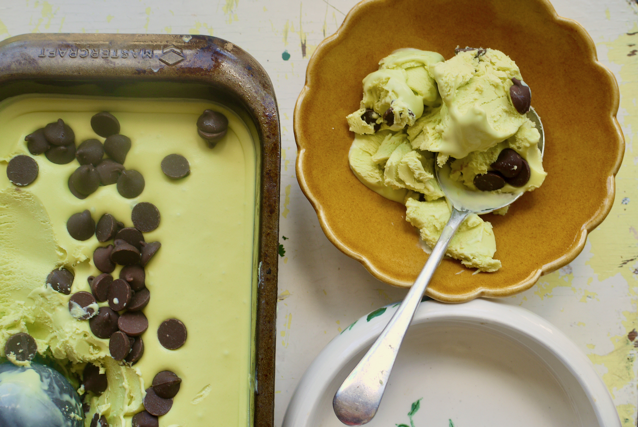 a brown fluted bowl of mint choc chip ice cream next to a tin of the ice cream with a spoon resting in the bowl
