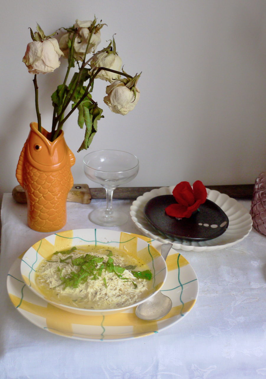 A bowl of soup on a white tablecloth with an orange vase of roses in the background