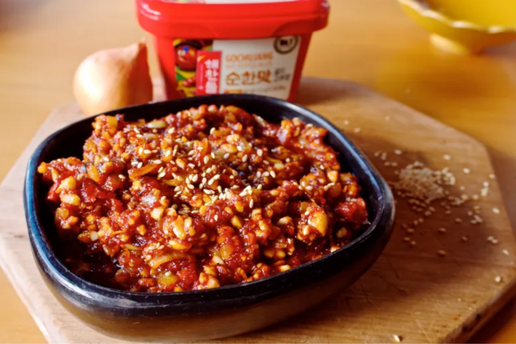 A bowl of gochujang bokkeum with a tub of gochujang and an onion behind it. This image links through to a web page with the recipe.