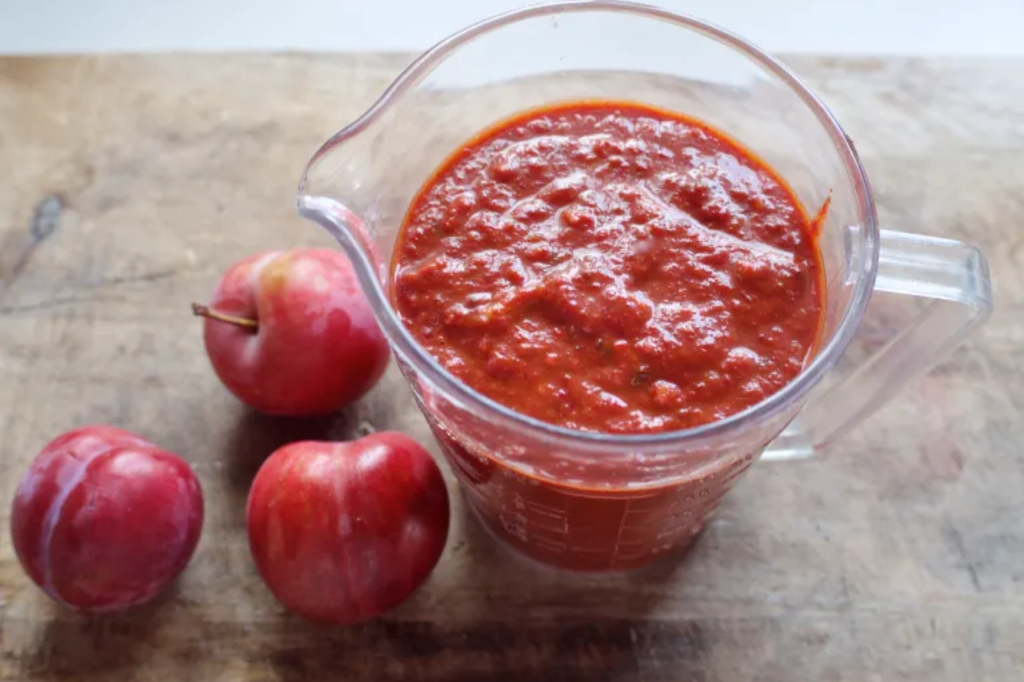 A jug of harissa with three plums on a wooden board. This image links through to a web page with the recipe.