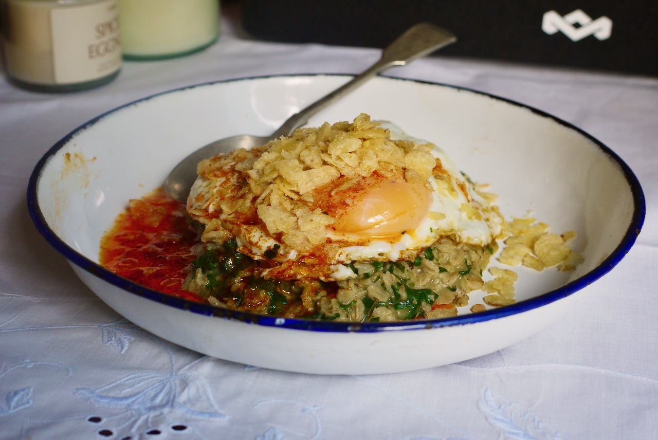 a white plate with a blue border and spoon resting on it with green oats and a fried egg, on a white tablecloth