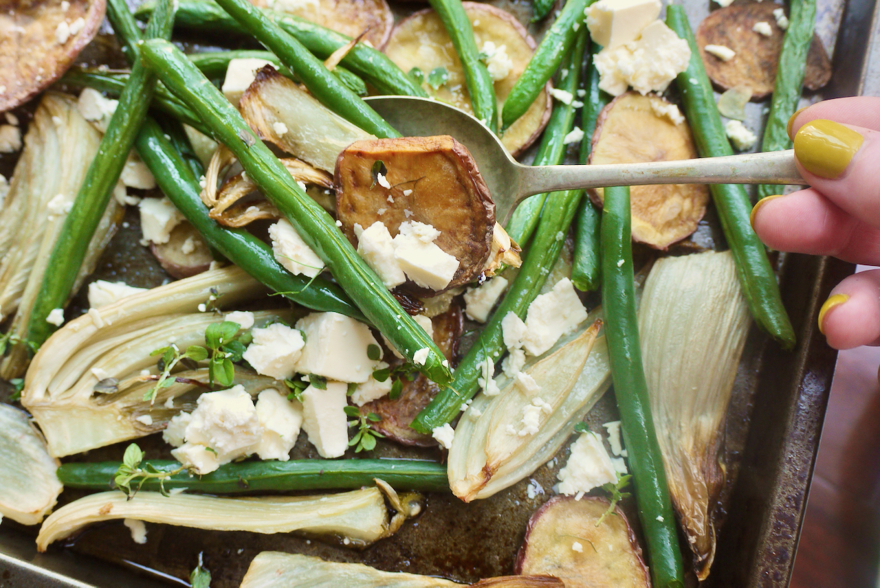 A hand with green fingernails holding a serving spoon of roasted potato, green beans, and feta