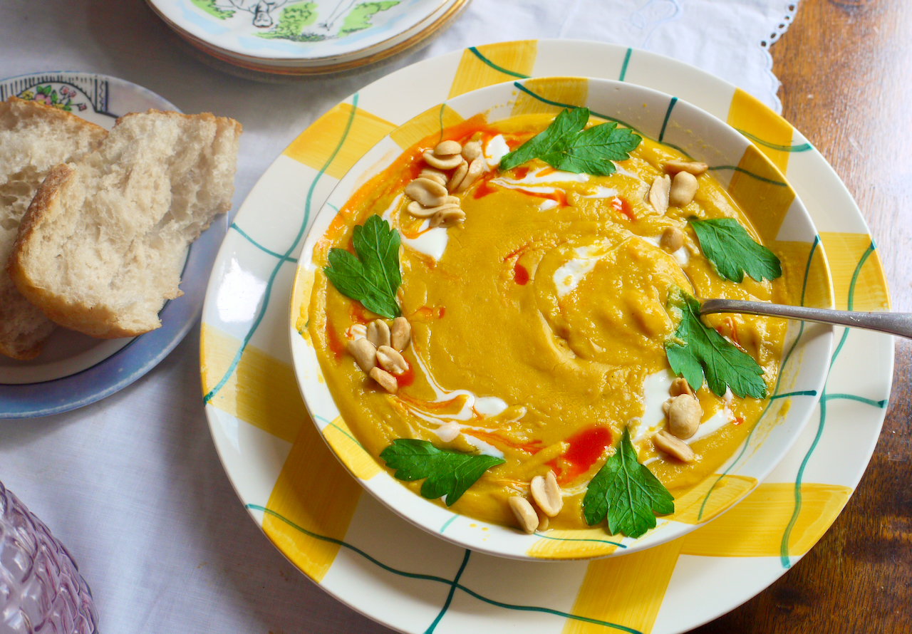 a spoon resting in a bowl of butternut soup with bread on a plate next to it