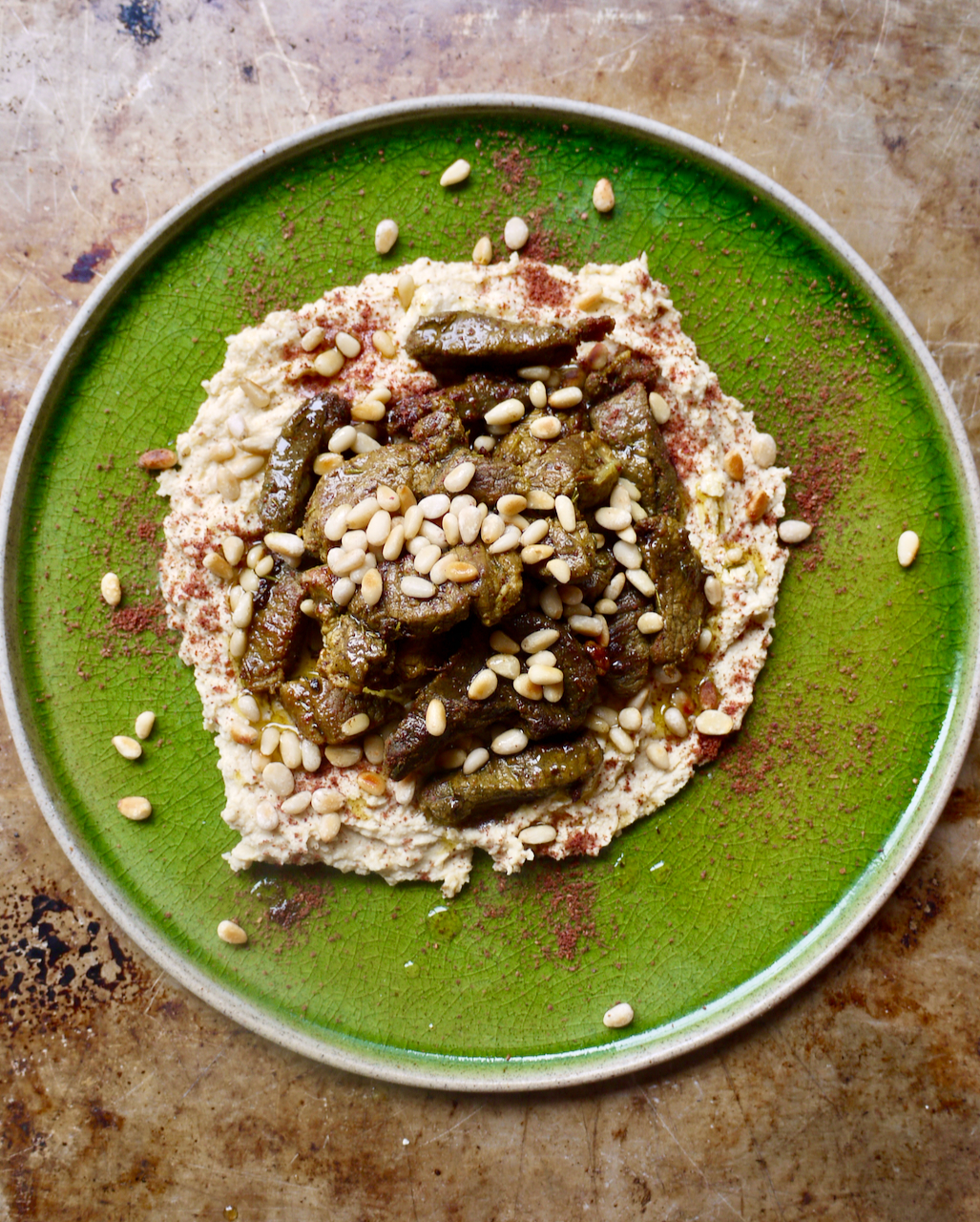 A green plate of hummus, lamb and pine nuts on a green plate, sitting on a baking tray