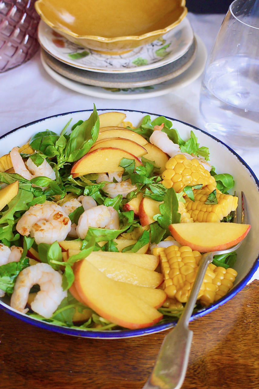 A blue-rimmed plate of corn prawn and peach salad with a stack of plates and water glass