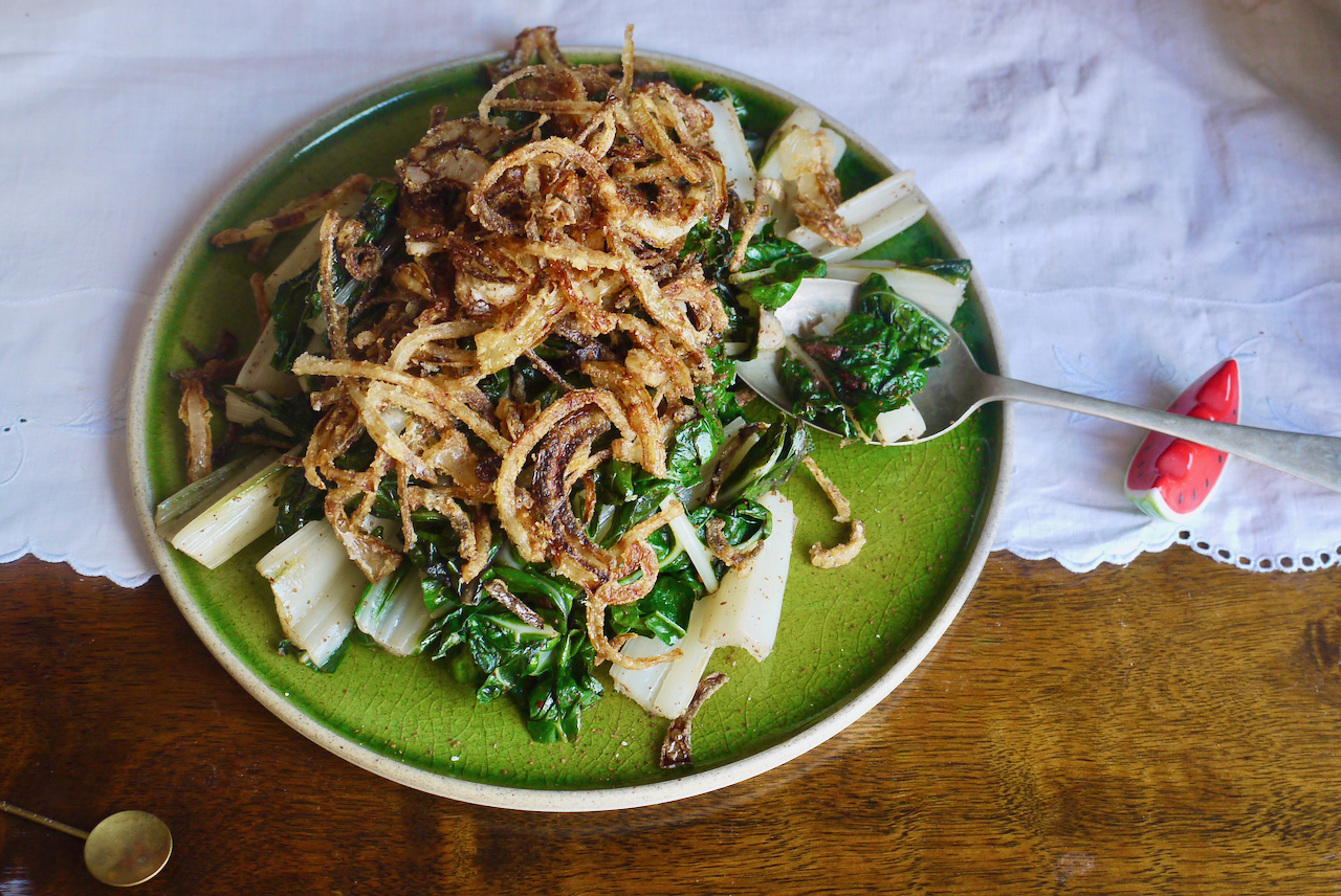 A serving spoon resting on a watermelon spoon rest with a green plate of braised silverbeet and fried onions