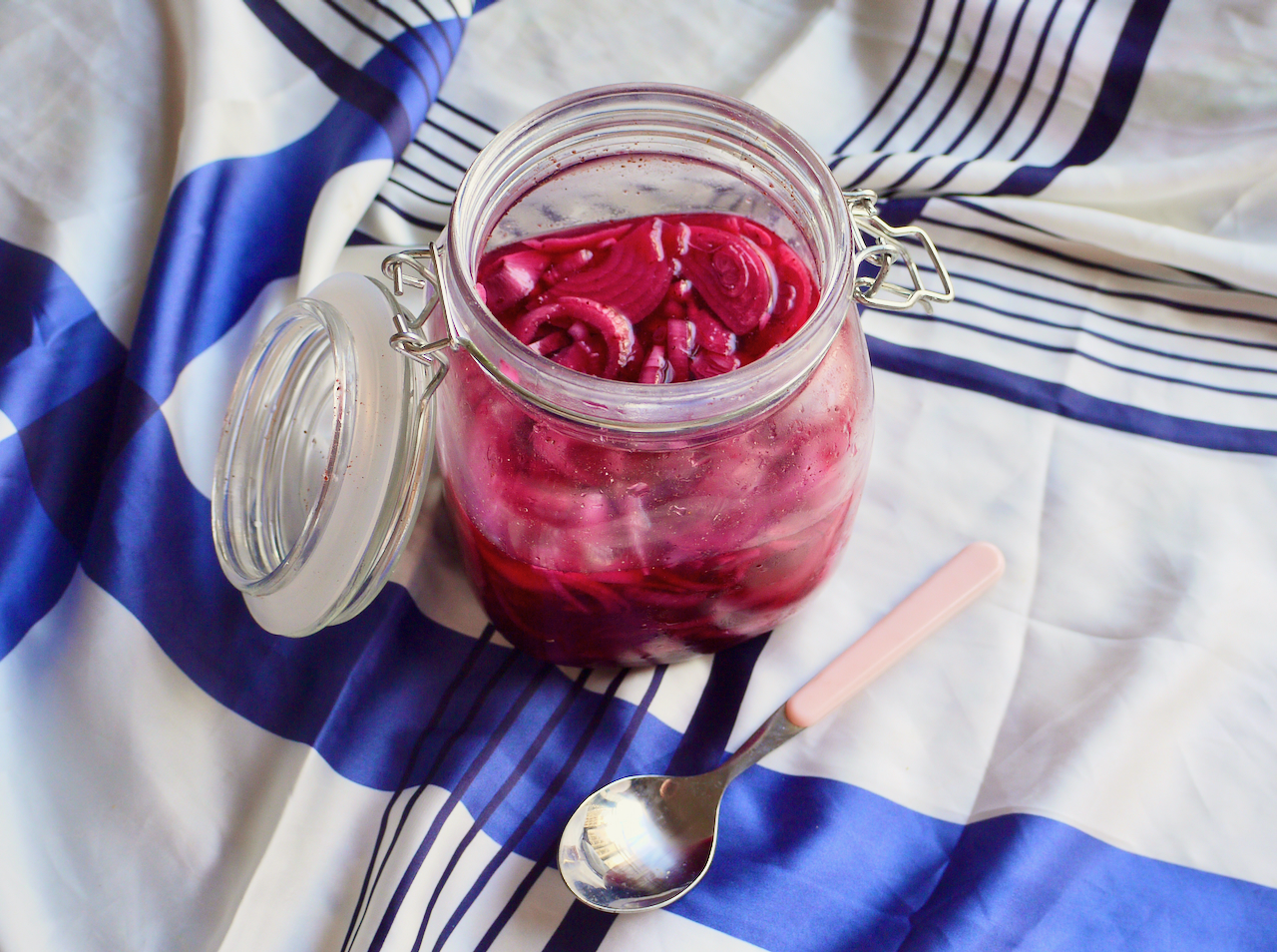 An open jar of pickled red onions on a blue and white cloth with a pink spoon