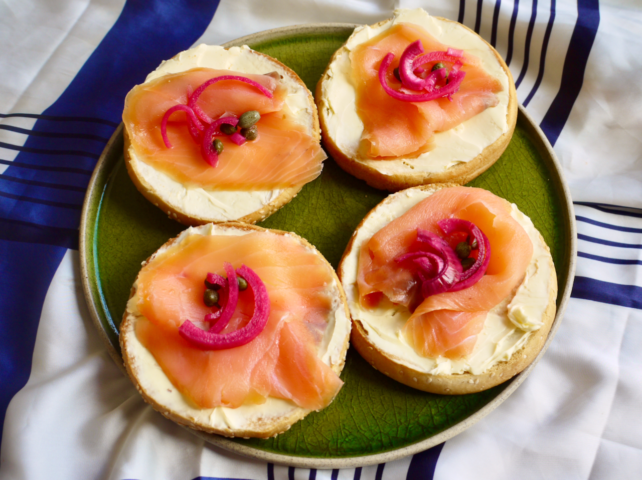 Four bagels with salmon and pickled red onions on a green plate on a blue and white cloth