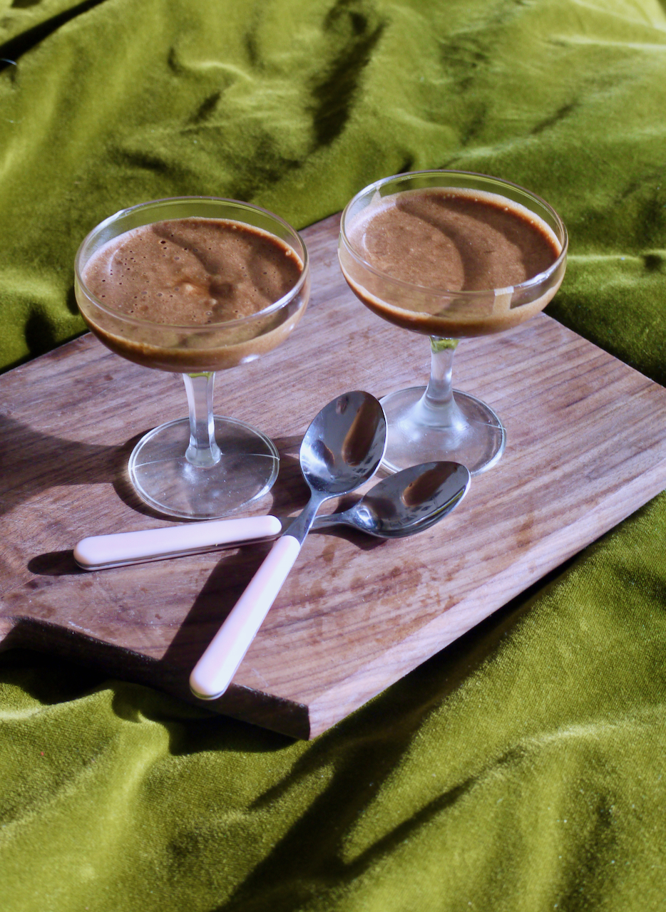 Two coupe glasses of chocolate mousse resting on a wooden chopping board with two teaspoons, on a vivid green velvet background