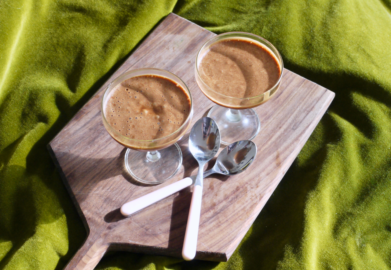 Two sunlit coupe glasses of chocolate mousse on a wooden board, on a green velvet background