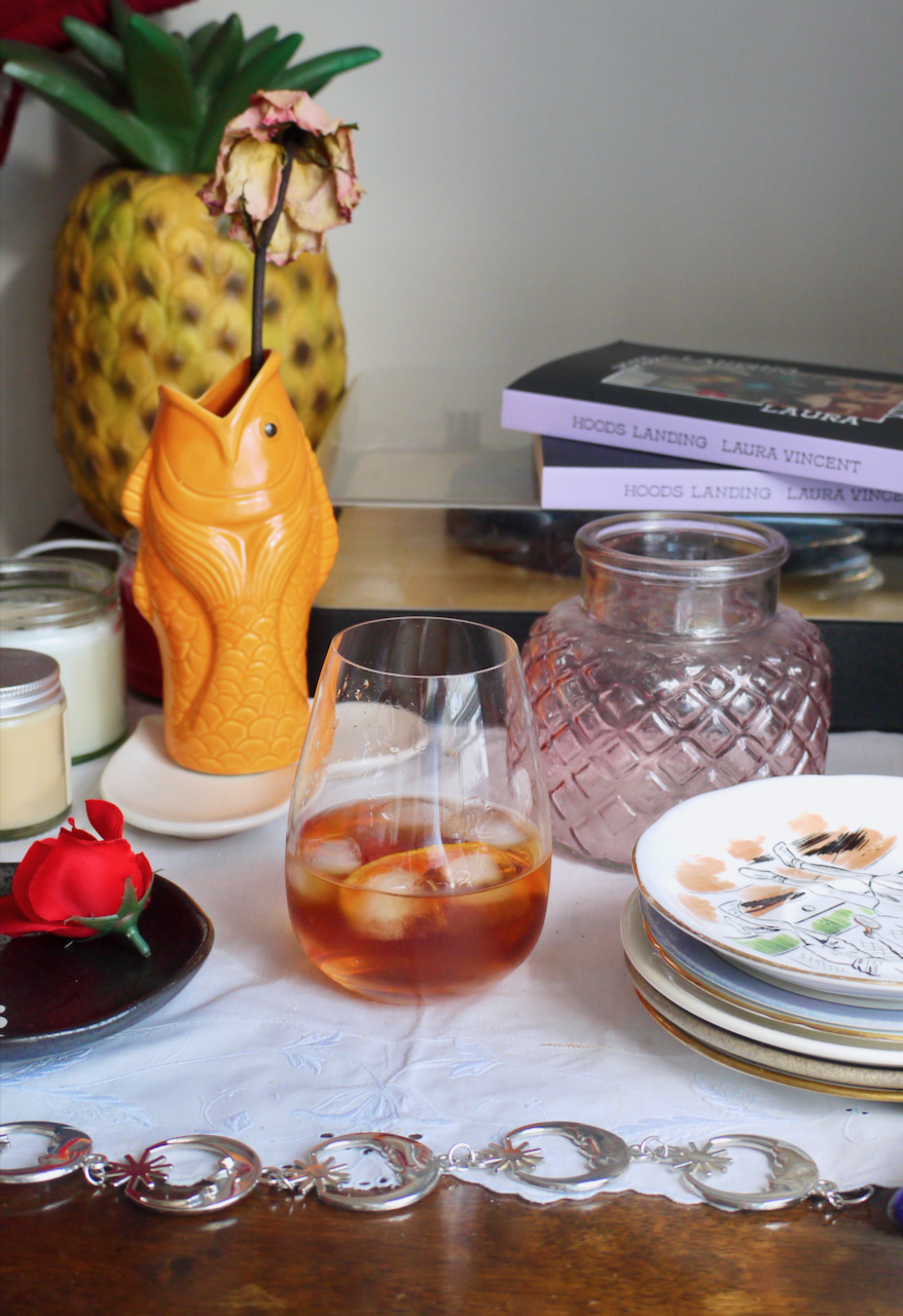 A cocktail in a glass with an orange peel garnish, with a goldfish vase and pineapple lamp behind it