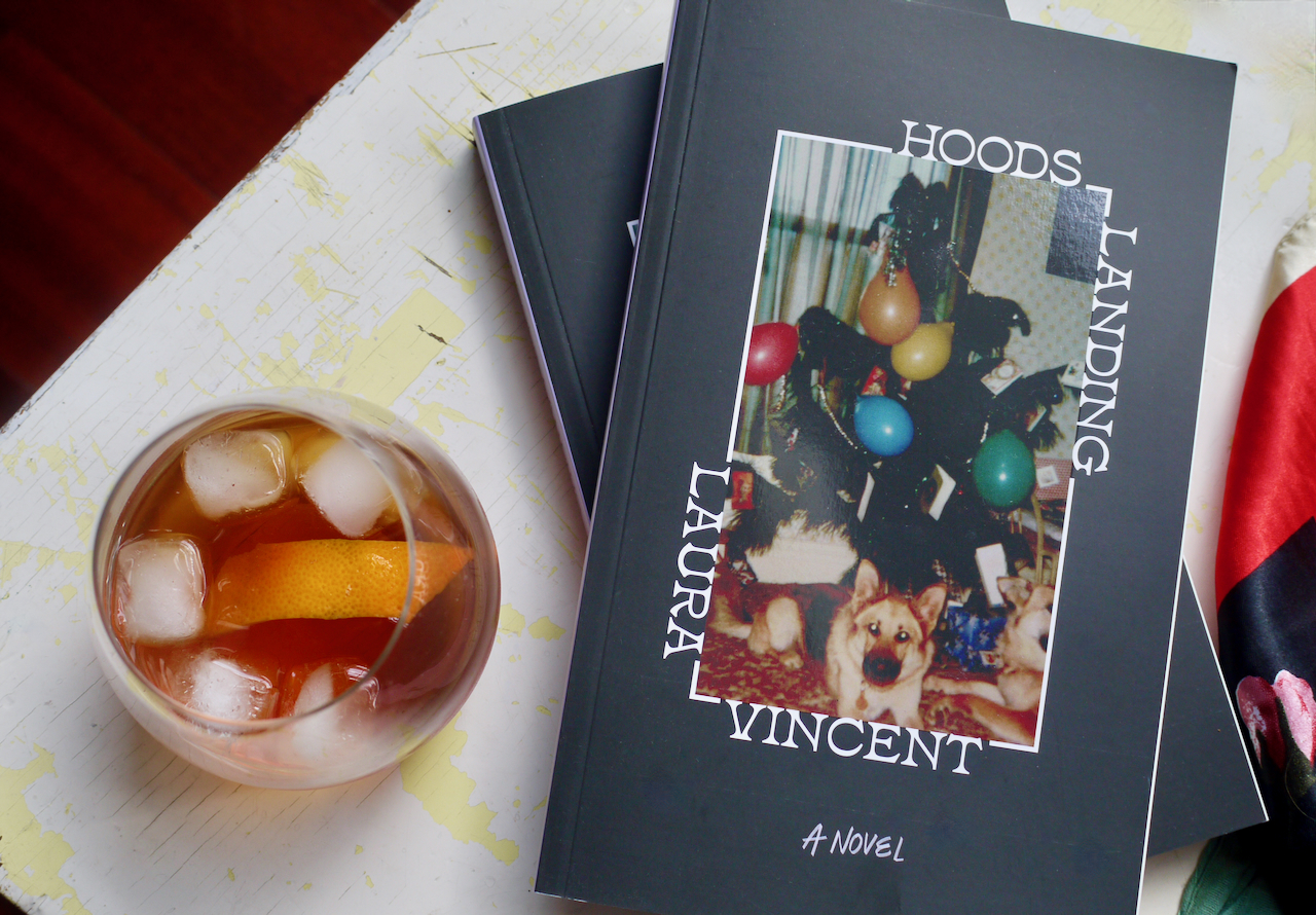 A cocktail with an orange peel garnish next to copes of Hoods Landing