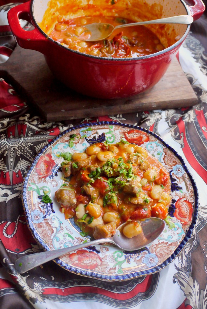 A blue and red plate of sausages and beans with a spoon resting on it, in front of a large saucepan of sausages and beans
