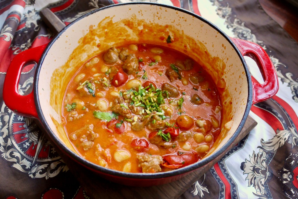 A spoon resting in a red saucepan of sausages and beans on a wooden board