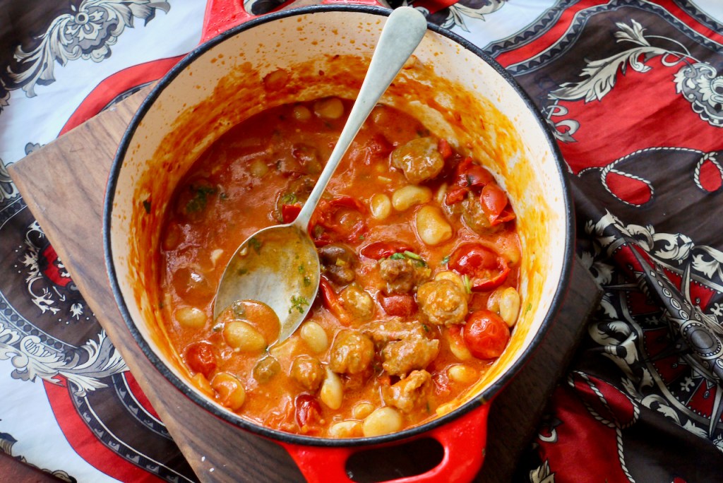 A red saucepan of sausages, beans and tomato with a spoon resting in it