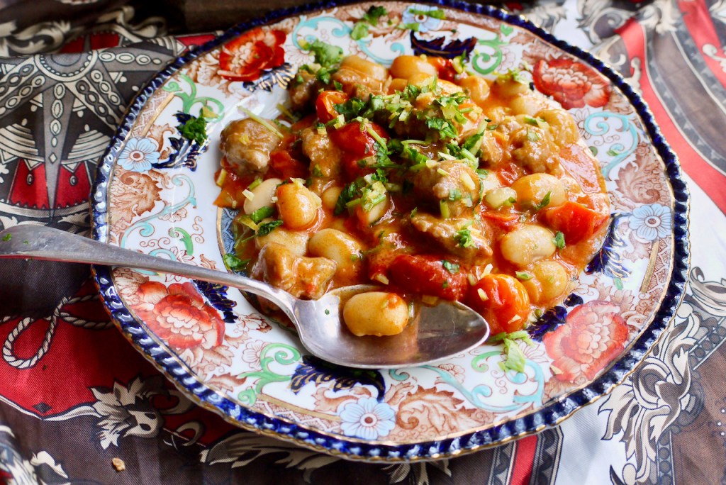 A spoon resting on a blue and red patterned plate of sausages and beans on a red and brown patterned cloth.jpeg