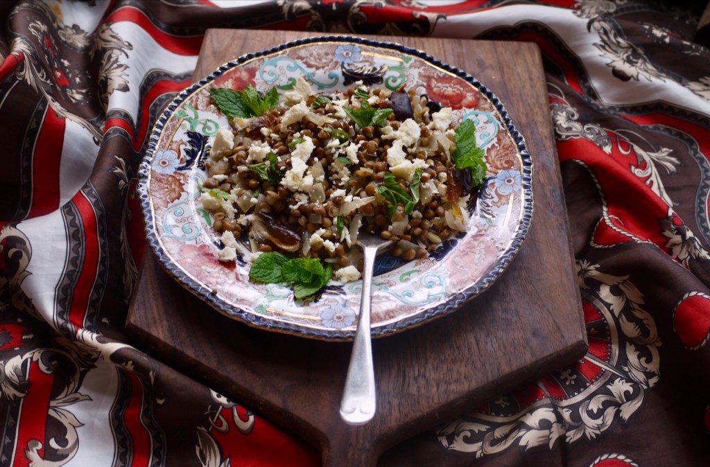 A blue and red plate of lentil salad with a fork on a wooden board on red and brown fabric