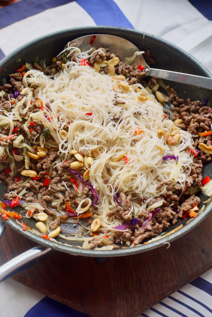 A pan of noodles and beef with a spoon on a wooden board on a blue and white cloth