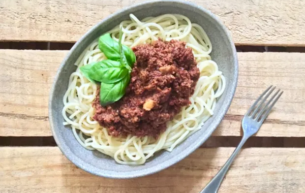 Vegan spaghetti bolognese in a grey oval bowl on a wooden table with a fork. This image links to the recipe.