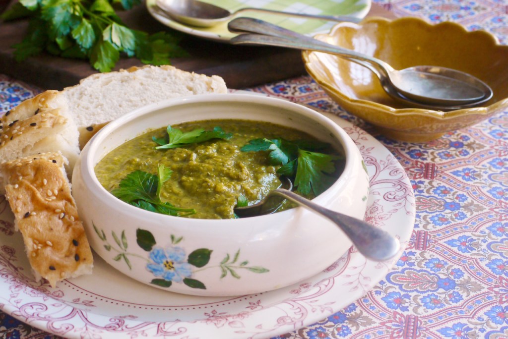 A bowl of soup sitting on a plate with several pieces of bread. In the background are plates with parsley and spoons on them