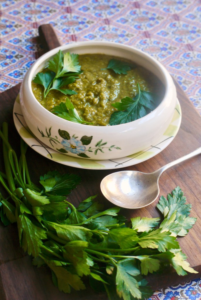 A bowl of soup on a wooden board with a bushel of flat leaf parsley and a spoon on it