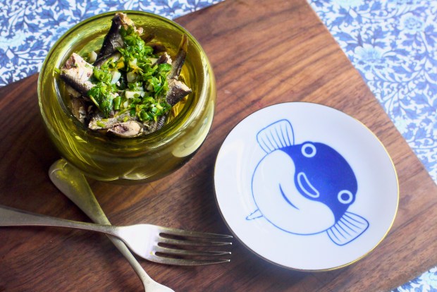 A green glass bowl of sardines and a small round plate with a blue fish on it, on a wooden board