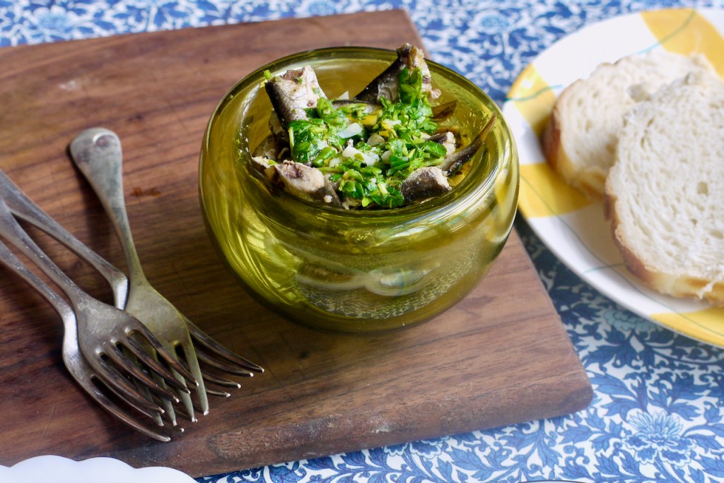 A green glass bowl of sardines on a wooden board with parsley sauce